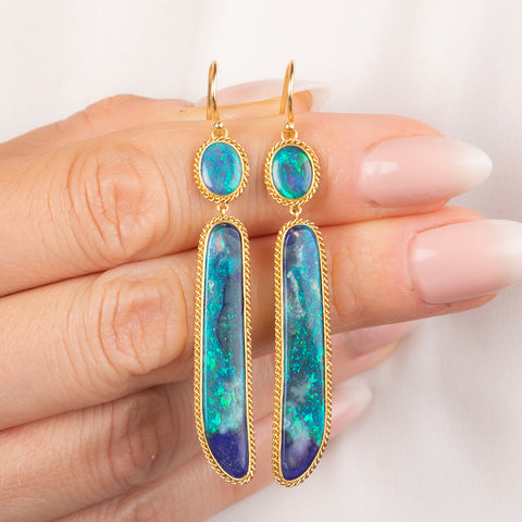 Close up of a gold dangle earring with oval and elongated black opal gemstones, held in a person's hand.