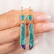 Close up of a gold dangle earring with oval and elongated black opal gemstones, held in a person's hand.