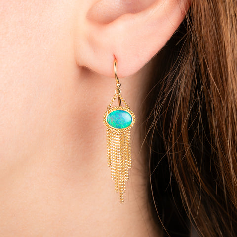 A pair of gold dangle earrings with oval blue-green opal stones and cascading gold fringe, shown worn by a model