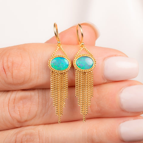 A pair of gold dangle earrings with oval blue-green opal stones and cascading gold fringe, shown held in a person's hand