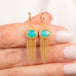 A pair of gold dangle earrings with oval blue-green opal stones and cascading gold fringe, shown held in a person's hand