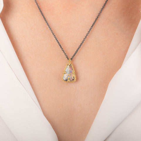 A necklace featuring a boulder opal set in an 18k gold bezel and suspended from an oxidized silver chain, worn on a person's neck.