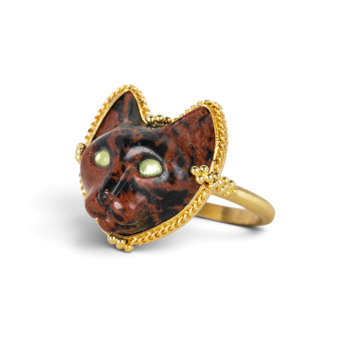 A gold ring featuring a carved dark brown and black cat face with green Tourmaline eyes, set against a clean white background.