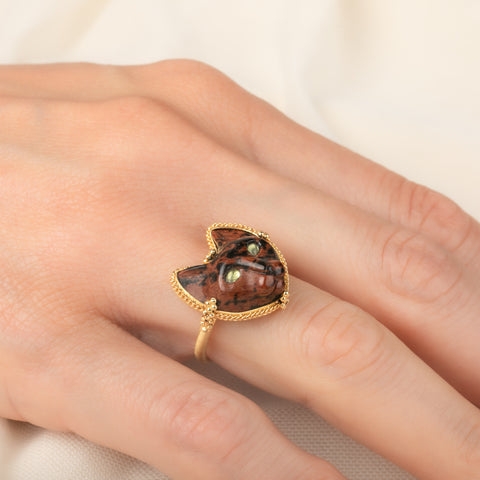 A gold ring featuring a carved dark brown and black cat face with green Tourmaline eyes, worn on a person's finger.