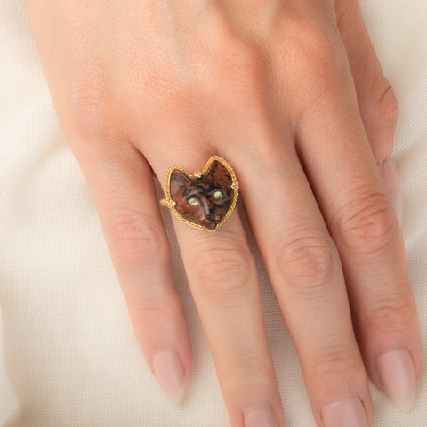 A gold ring featuring a carved dark brown and black cat face with green Tourmaline eyes, worn on a person's finger.