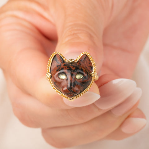 A gold ring featuring a carved dark brown and black cat face with green Tourmaline eyes, held in hand.