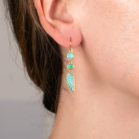 A model wears a handmade gold earring featuring a blend of emerald and opal gemstones, that is punctuated with a hand-carved wing stone.