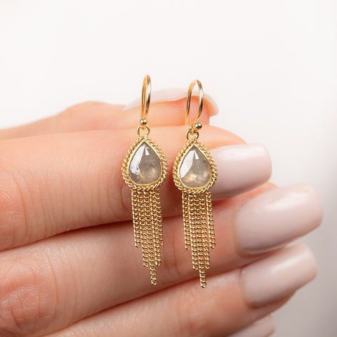 A pair of gold dangle earrings with teardrop-shaped diamonds and delicate gold chain fringe, held between fingers against a clean white background.