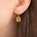 A close-up of a person's ear wearing a gold drop earring with a rectangular citrine gemstone in a textured bezel setting.