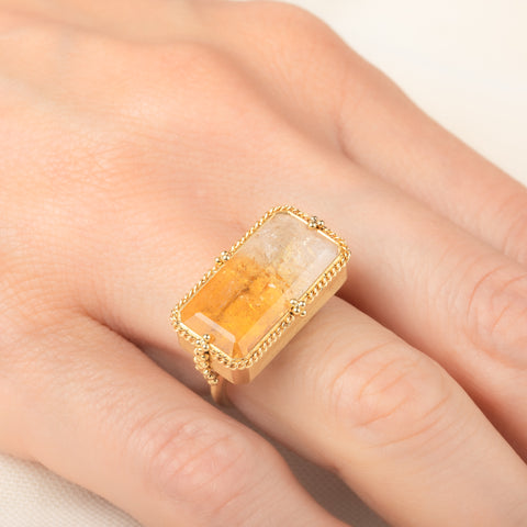 A gold ring featuring a large, rectangular, Citrine gemstone set in a detailed gold bezel, worn on a person's finger.
