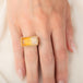 A gold ring featuring a large, rectangular, Citrine gemstone set in a detailed gold bezel, worn on a person's finger.