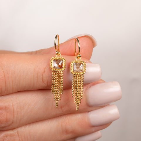 A pair of gold dangle earrings with square diamonds and gold chain tassels, held in hand.