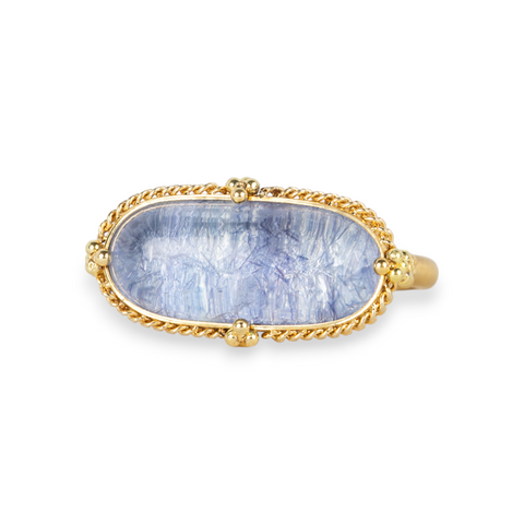 An oval ring featuring a textured light blue dumortierite stone in a yellow gold bezel setting and a matching gold band, shown on a white background.