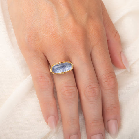 An oval ring featuring a textured light blue dumortierite stone in a yellow gold bezel setting and a matching gold band, worn on a person's finger.