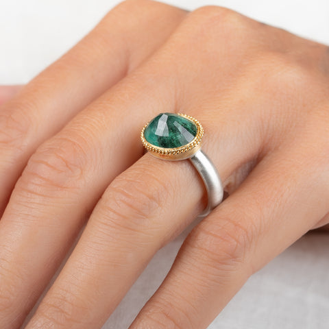 An oval emerald gemstone ring set in a gold bezel, with a sterling silver band, on a finger
