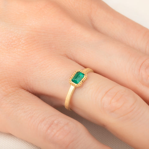 A yellow gold ring featuring a rectangular emerald gemstone in an 18k gold bezel setting, worn on a person's finger.
