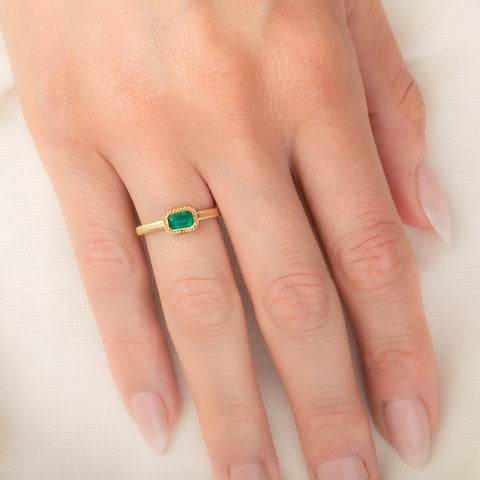 A yellow gold ring featuring a rectangular emerald gemstone in an 18k gold bezel setting, worn on a person's finger.