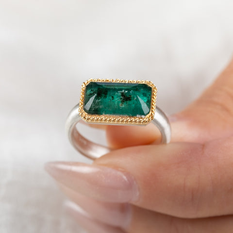 A rectangular emerald stone ring with a gold bezel and a sterling silver band, held in hand