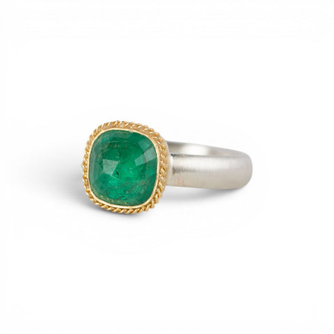 A ring featuring a cushion-cut emerald gemstone in a gold bezel setting, paired with a silver band, shown against a plain white background.