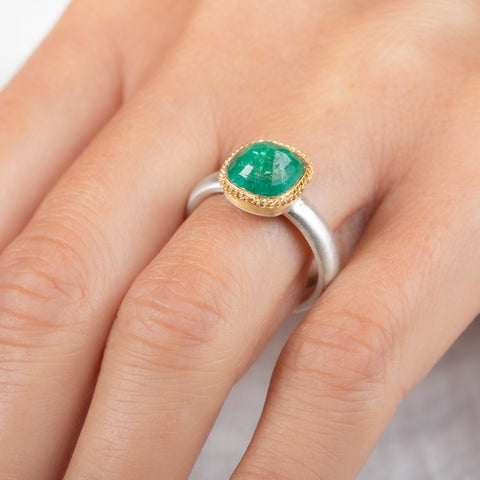A ring featuring a cushion-cut emerald gemstone in a gold bezel setting, paired with a silver band, worn on a hand