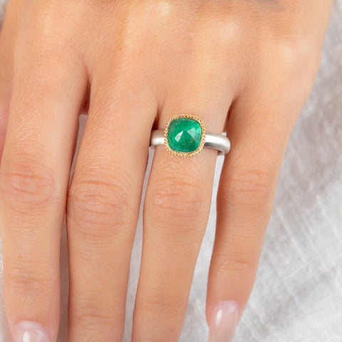 A ring featuring a cushion-cut emerald gemstone in a gold bezel setting, paired with a silver band, worn on a hand
