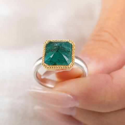 An emerald gemstone ring with a gold bezel and sterling silver band, held in hand