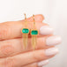 A pair of gold dangle earrings with rectangular emerald gemstones and gold chain fringe, held in a person's hand.