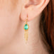 Gold earrings featuring emerald gemstones and gold chain tassels, shown  worn in a person's ear.