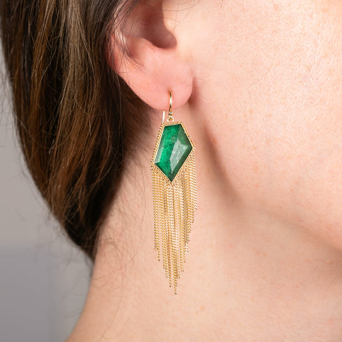 An emerald gemstone earring with gold fringe worn by a person against a neutral background.