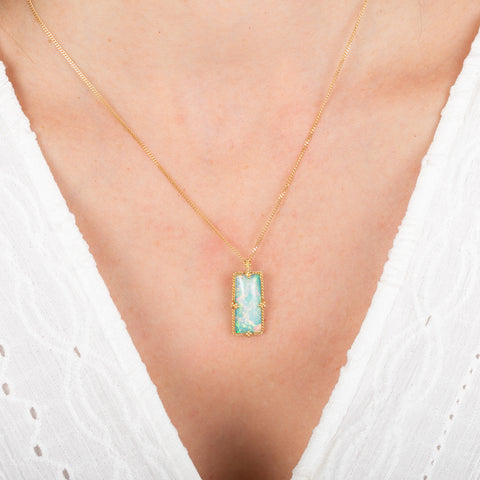 Gold necklace with a rectangular Ethiopian opal pendant on a person wearing a white top.
