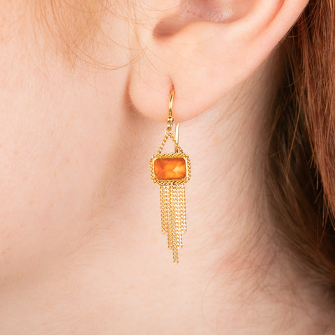 Gold earrings featuring fire opal gemstones and gold chain tassels, shown worn on a model's ear.