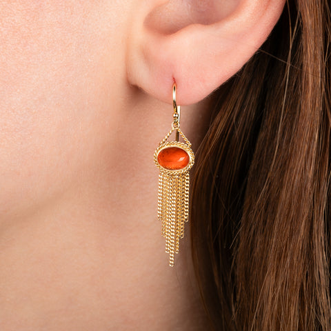 A gold dangle earring with a fire opal orange stone, shown worn on a person's ear with brown hair visible.