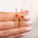 A pair of gold dangle earrings with fire opal orange gemstones and cascading chain fringe, held between fingers against a soft white background.