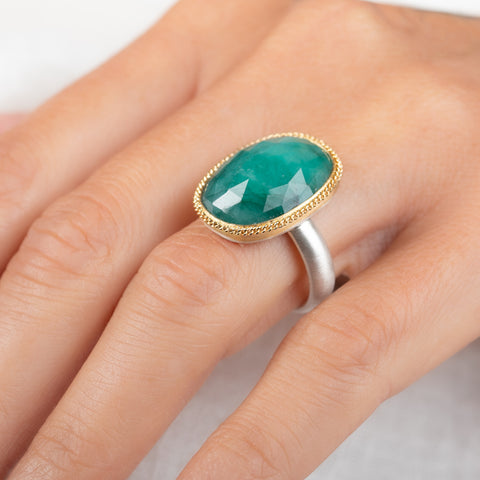 An oval emerald gemstone ring with a textured gold bezel and a sterling silver band worn on a person's finger