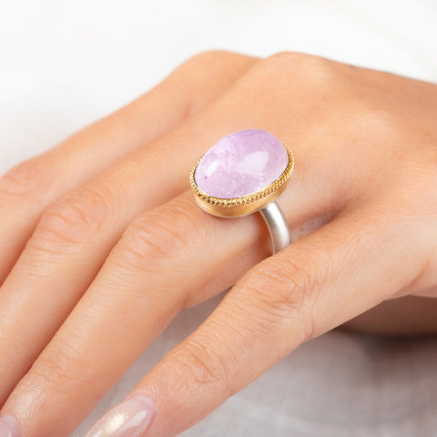 A ring featuring a large oval kunzite stone, set in a gold textured bezel with a sterling silver band, worn on a person's finger.