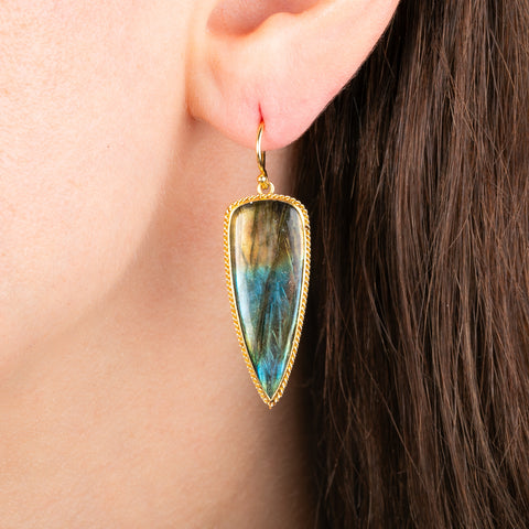 A close-up of a person's ear wearing a gold dangle earring with a long teardrop-shaped iridescent labradorite stone.
