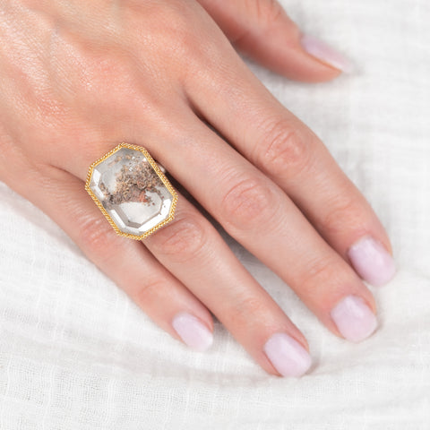 Hand wearing a gold and silver ring with a lodolite quartz stone on a white background