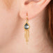 Gold earring featuring London blue topaz teardrop-shaped gemstone, and gold chain tassel, shown worn on a person's ear.