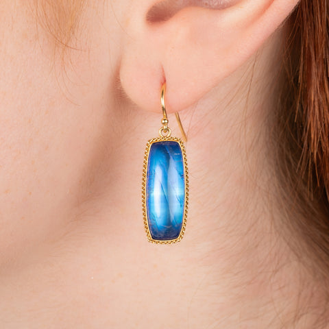 Gold earrings featuring large, blue moonstones, worn in a model's ear.