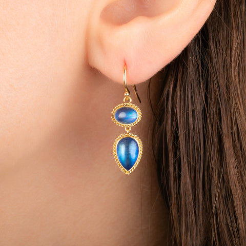 A close-up of a person's ear wearing a gold drop earring with two blue moonstones.