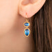 A close-up of a person's ear wearing a gold drop earring with two blue moonstones.