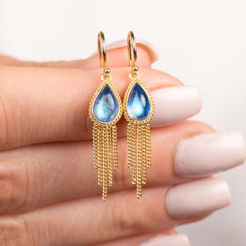 A pair of gold dangle earrings with teardrop-shaped moonstones and gold chain fringes, held between fingers with light pink manicured nails against a soft white background.