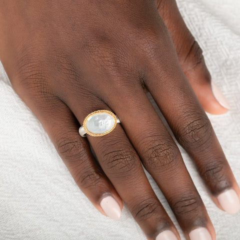 Gold and silver ring with a mother of pearl on a person's finger against a neutral background