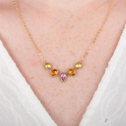 A gold necklace featuring a row of sapphire gemstones in different shapes and colors, shown worn on a model's neck.