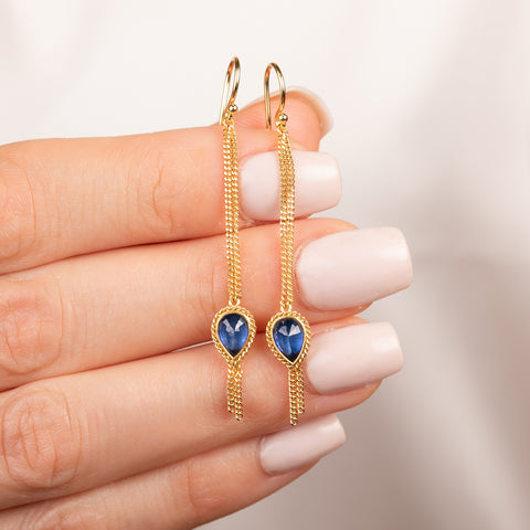 A pair of gold drop earrings with teardrop-shaped Sapphire gemstones and dangling chain details, held in hand.
