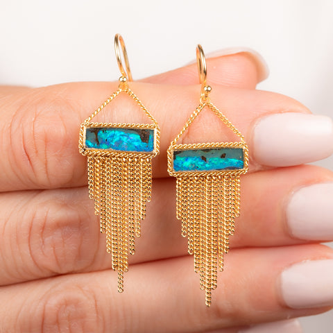 A pair of gold dangle earrings featuring a rectangular boulder opal and cascading gold chain fringe, shown held in hand.