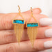A pair of gold dangle earrings featuring a rectangular boulder opal and cascading gold chain fringe, shown held in hand.