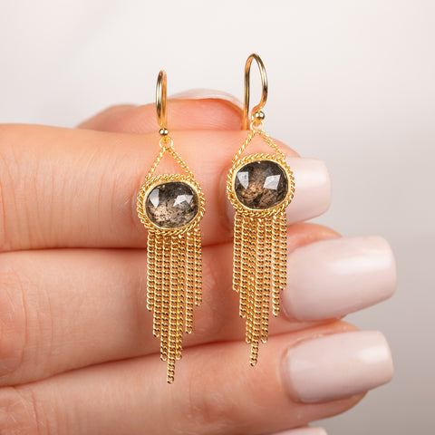 A pair of gold dangle earrings with oval diamond gemstones and cascading gold chain fringe, show held in hand.