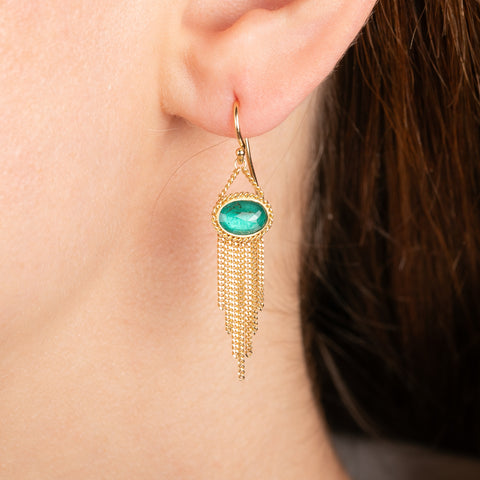 A pair of gold dangle earrings with oval emerald gemstones and cascading gold chain fringe, shown worn in a model's ear.