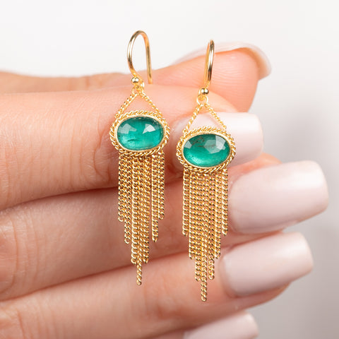 A pair of gold dangle earrings with oval emerald gemstones and cascading gold chain fringe, shown held in hand.